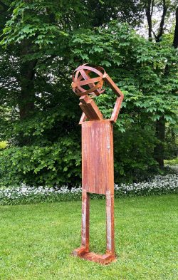 Abstrakte, anthropomorphe Skulptur aus rostpatiniertem Stahl, die in reduzierter Linienführung eine stilisierte Kugel auf den Schultern trägt.