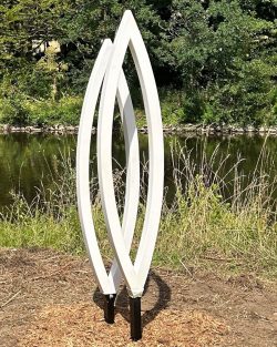 Zwei vertikale, linsenförmige Rahmenskulpturen in Weiß an einem Flussufer vor grünem Naturhintergrund.
