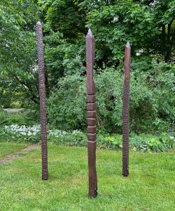 Skulpturengruppe aus drei schlanken, dunkel gemusterten Holzstelen mit strukturierten Oberflächen auf einer Rasenfläche.