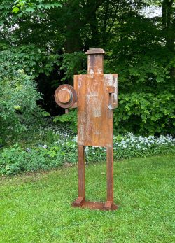 Abstrakte anthropomorphe Skulptur aus rostpatiniertem Stahl mit Hammer- und Schild-Symbolik auf einer Rasenfläche vor Bäumen.