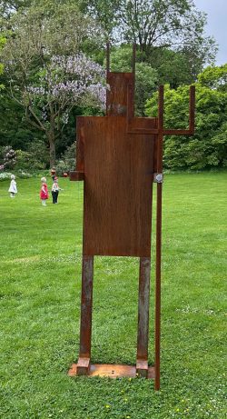 Abstrakte anthropomorphe Skulptur aus rostigem Stahl mit Dreizack-Symbolik auf einer Rasenfläche in einer Parkanlage.