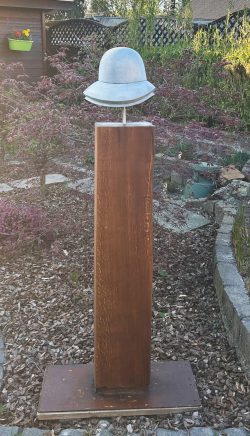 Moderne Skulptur einer helmartigen Kopffigur aus hellem Metall auf einer hohen, massiven Holzstele in einem Garten.