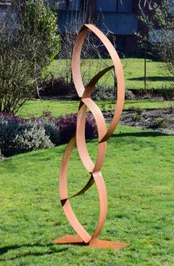 Abstrakte, vertikale Skulptur aus in sich verdrehten und verschlungenen Metallbändern mit Rost-Patina auf einem Rasen.