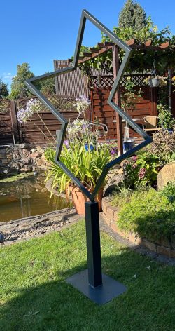 Abstrakte, geometrische Rahmenskulptur aus dunklem Profilstahl, die den Blick auf eine Gartenlandschaft mit Teich freigibt.