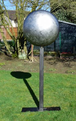 Abstrakte Skulptur einer großen Stahllinse auf einer hohen, schlanken Stele in einer Gartenanlage.