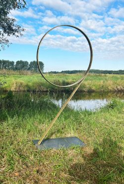 Abstrakte, dynamisch geschwungene Metallskulptur in Form einer Schleife auf einer Schieferplatte in einer Flusslandschaft.