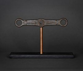 Symmetrische, minimalistische Skulptur aus einem horizontalen, doppeläugigen Metallsteg auf einem dünnen Stab und dunkler Basis.
