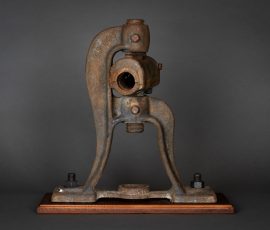 Abstrakte Assemblage aus einem geschwungenen, industriellen Formteil aus Metall auf einem profilierten Holzsockel.