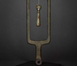 ertikale, gabelförmige Metallskulptur mit einem im Zentrum frei schwingenden Pendel- oder Senklot-Element.