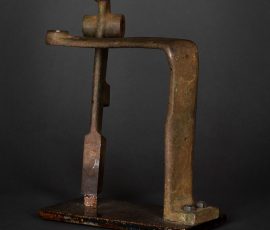 Abstrakte Skulptur aus einem massiven, rechtwinklig gebogenen Metallelement mit industrieller Patina auf einer dunklen Basis.