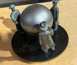 Konzeptuelle Skulptur auf einer runden Basis, bei der sich stilisierte Figuren um eine zentrale, matte Metallkugel gruppieren.