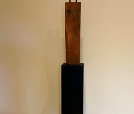 Schlichte, minimalistische Skulptur bestehend aus einem naturbelassenen, vertikalen Holzblock auf einer schlanken, schwarzen Stele.